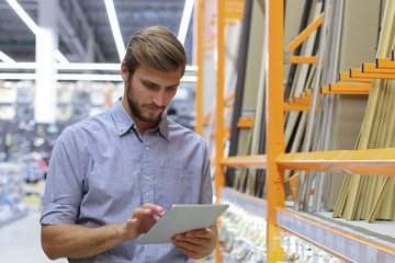 Young man shopping or working in a hardware warehouse standing checking supplies on his tablet.