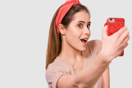 Closeup portrait of beautiful young European woman in casual t-shirt and red head band, taking self portrait over white studio wall. Happy amazed female smiling and taking a selfie on her smart phone.