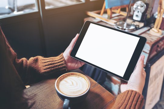 Mockup image of hands holding black tablet pc with blank white screen with coffee cup on wooden table