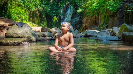 Young boy sitting in a stream enjoying the water amd rainforest around him.