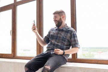 Cute young positive guy hipster with a beard is sitting on the windowsill by the big window and talking on the video communication using a smartphone and wireless high-speed Internet.