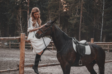 Beautiful and elegant young woman wearing the dress is stroking the horse on the ranch. An attractive rider is posing outdoors near the saddle. Forest, nature landscape, countryside