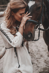 Beautiful and ladylike young woman wearing the dress is embracing and stroking the horse on the ranch. An attractive rider is posing outdoors near the saddle. Summertime, nature landscape, countryside