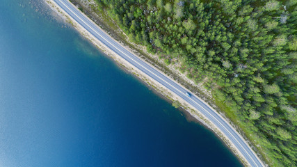 Aerial view of road between green summer forest and blue lake. Blue car  moving on road.