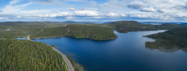 Aerial drone view of a road through forest and hills in Lapland on a cloudy day. Beautiful lake with blue water. 