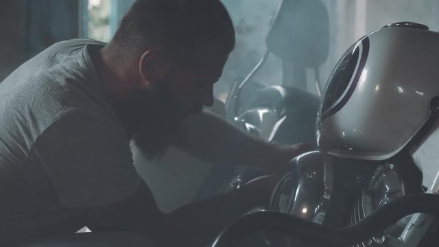 biker with a motorcycle in an atmospheric garage. a bright-looking man with a beard and tattoos is preparing his iron horse for the ride.