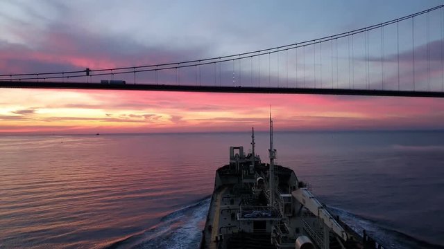 bulk carrier with steel towers sails near huge bridge with driving cars silhouettes at summer sunset