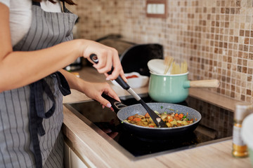 Woman in apron standing next to stove in kitchen and cooking healthy dinner