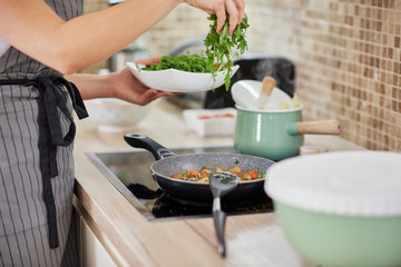 Woman in apron standing next to stove in kitchen and putting rucola in the meal