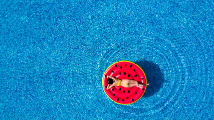 Aerial top view of woman wearing a hat sitting in inflatable watermelon in calm waters of a pool