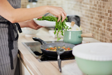 Woman in apron standing next to stove in kitchen and putting rucola in the meal
