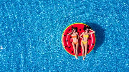 Aerial view of two women lying on inflatable watermelon mattrass floating and relaxing in swimming pool