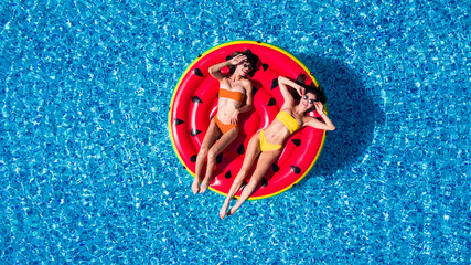 Aerial view of two women lying on inflatable watermelon mattrass floating and relaxing in swimming pool