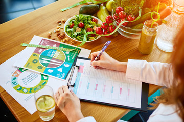 Woman dietitian in medical uniform with tape measure working on a diet plan sitting with different healthy food ingredients in the green office on background. Weight loss and right nutrition concept