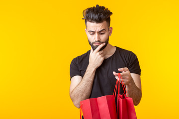 Cute young sad bearded man looks at the purchase in shopping bags on a yellow background with copy space. Concept of rash and waste of money.