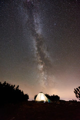 Tourist camping at summer night on the top of rocky mountain. Glowing tent under incredibly beautiful night sky full of stars and Milky way. On background amazing starry sky and mountains