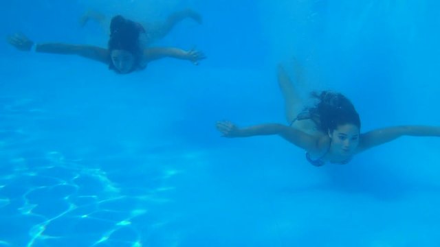 young women friends into bathing suit dives and floating underwater in clear blue swimming-pool during summer leave at resort, underwater shooting view