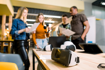 Blurred image of five multiethnical business persons are developing a project using modern gadgets and virtual reality goggles. Focus on the VR goggles on the table. technologies of the future