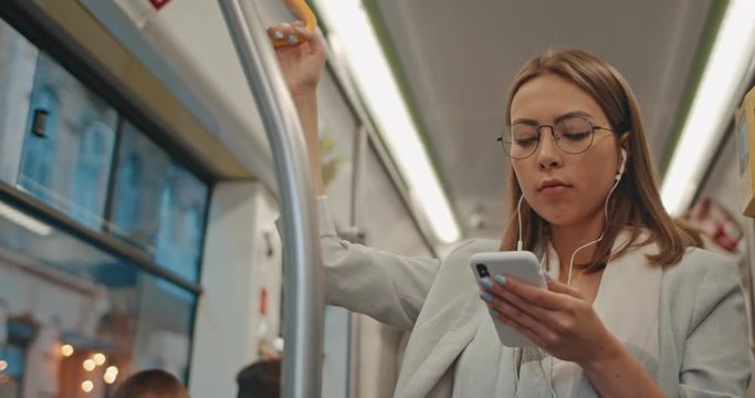 Portrait of cute brunette girl in glasses and headphones holds the handrail, listening to music and browsing on mobile phone in public transport.