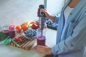 Woman cooking fresh blueberry smoothie with hand blender at kitchen at home. Healthy eating and berry drink