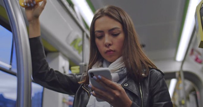 Portrait of cute brunette woman holds the handrail, texting and browsing on mobile phone in public transport. City lights background.