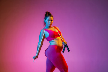Young caucasian plus size female model's training on gradient purple background in neon light. Doing workout exercises with the jump rope. Concept of sport, healthy lifestyle, body positive, equality.