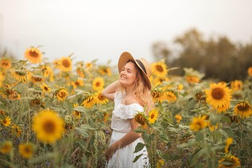 Naklejka na ściany i meble Beautiful curly young woman in a sunflower field holding a wicker hat. Portrait of a young woman in the sun. Summer.