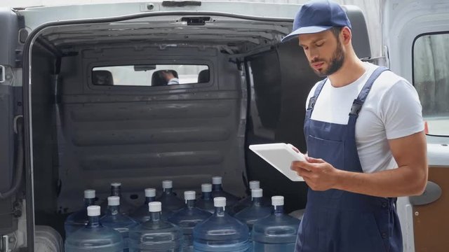 handsome bearded man in cap using digital tablet while standing near car with bottles of water 