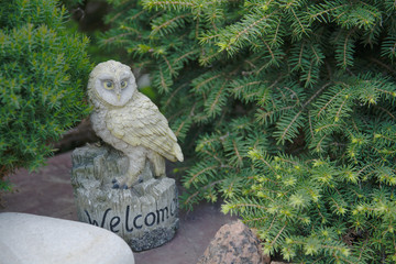 A garden owl figurine with a greeting standing on a large stone against a background of green fir...
