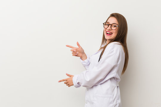 Young Doctor Woman Against A White Wall Excited Pointing With Forefingers Away.