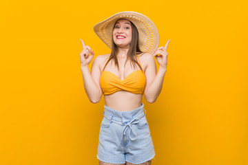 Young caucasian woman wearing a straw hat, summer look indicates with both fore fingers up showing a blank space.