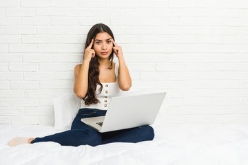 Young arab woman working with her laptop on the bed focused on a task, keeping forefingers pointing head.