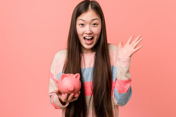 Young asian woman holding a piggy bank celebrating a victory or success