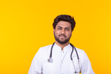 Positive young Indian doctor in a white coat with a stethoscope posing on a yellow background. Concept of effective medicine and doctor's advice.