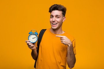 Young student man holding an alarm clock pointing with finger at you as if inviting come closer.