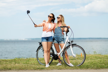 leisure, technology and friendship concept - happy smiling teenage girls or friends with bicycle taking picture by selfie stick on smartphone at seaside in summer