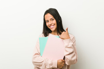 Young hispanic woman holding some notebooks smiling and raising thumb up