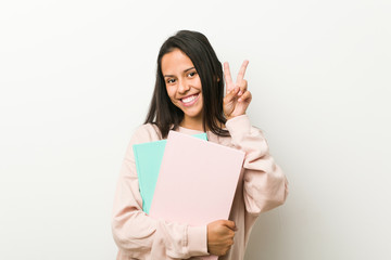 Young hispanic woman holding some notebooks showing victory sign and smiling broadly.