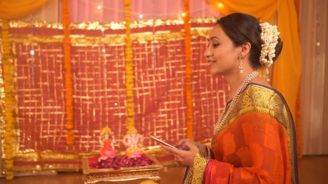 Attractive Indian woman texting using a smartphone before pooja begins at home - Festive Background. Indian stock footage of Indian woman standing in mandir / temple and texting on her phone happil...