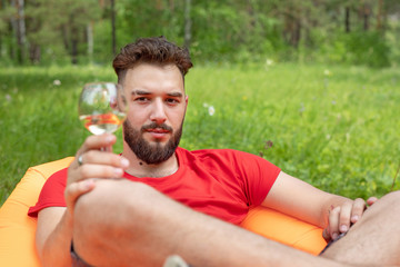 Leisure concept, camping, summer vacation. Young bearded caucasian man with glasses of wine laying at orange sofa. Relaxing and enjoying life on air bed.