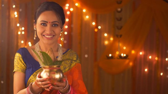 Indian traditional woman holding a copper pot / kalash with coconut and mango leaves for traditional customs. Indian stock video of young woman decorating house and pooja ghar with traditional elem...