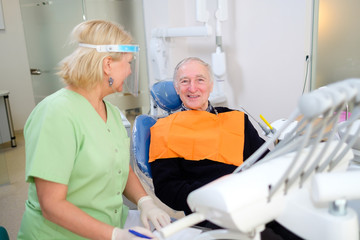 Satisfied senior male patient is smiling while sitting in stomatologic armchair next to positive female dentist who is smiling and standing nearby. Concept of accessibility of medicine for seniors