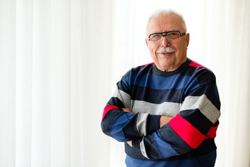 Portrait of smiling and confident with cross arms senior man 70-75 years old with eyeglasses stands near light window. Text space for ad