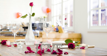 Obraz na płótnie Canvas beautiful romantic set up dinner table at home with flowers and rose petals surrounding love letter on wooden desk on valentine day. kitchen in background. candles warm up the room with light up fire