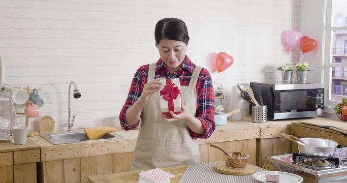 slow motion cute smiling young asian woman opening gift box in wooden modern kitchen on valentine's day. girl cheerful recieved surprised with love. card letter on table while making dessert.