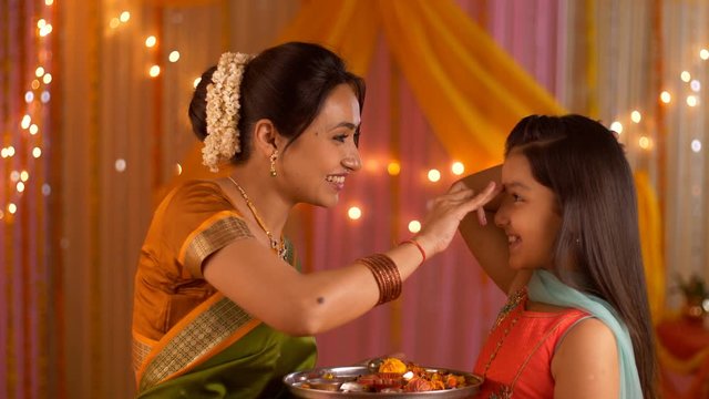 Mother putting tilak on young daughter's forehead - Celebration at home. Festive colorful background. Indian stock footage of nuclear family celebrating Diwali festival at home - Mother-daughter pe...