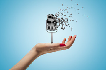 Female hand holding vintage microphone shattering into pieces on blue background