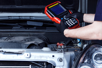 Closeup mechanic hands are holding special diagnostic equipment, scanner at vehicle with open hood. Repairman is repairing car and detecting problems at service station car repair shop.