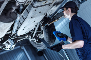 Mechanic is repairing car at service station. Repairman is detecting problems by special software on laptop and diagnostic tool at workshop auto repair shop. Vehicle on hydraulic lift is above.