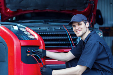 Mechanic is repairing car at service station. Repairer in jumpsuit is conducting diagnostics. Vehicle is connected to equipment for maintenance and refueling of air conditioners in auto repair shop.